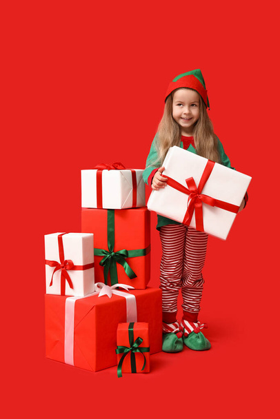 Nettes kleines Mädchen im Weihnachtselfenkostüm mit passenden Schachteln auf rotem Hintergrund - Foto, Bild