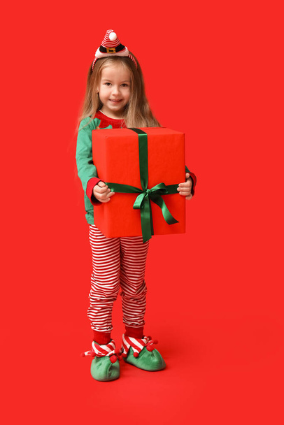 Cute little girl in Christmas elf costume with gift box on red background - Fotoğraf, Görsel