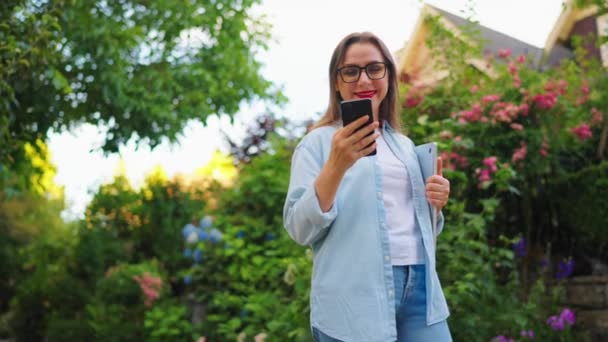 Boldog nő laptoppal és okostelefonnal a kezében áll a virágzó kertben vagy a zöld utcában munka után nap. Lassú mozgás. - Felvétel, videó