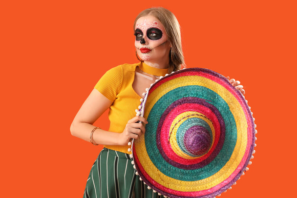 Nuori nainen sombrero hattu ja maalattu kallo hänen kasvonsa oranssi tausta. Meksikon kuolleiden päivän juhla (El Dia de Muertos) - Valokuva, kuva