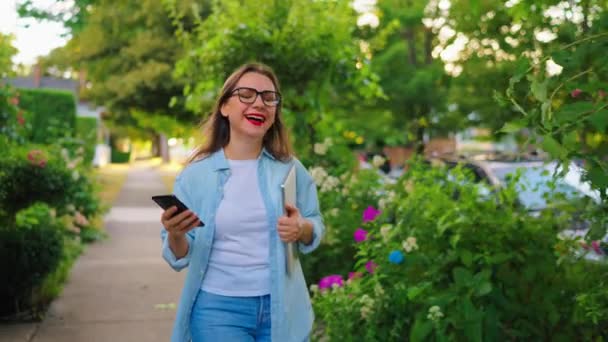 Щаслива жінка з ноутбуком і смартфоном в руках ходить крізь квітучий сад або зелену вулицю після робочого дня. Повільний рух - Кадри, відео