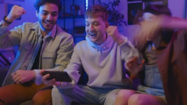 Μέτριο πλάνο της νεαρής Καυκάσιας γυναίκας και δύο διαφορετικών ανδρών κάθονται μαζί στον καναπέ στο σπίτι με smartphone, βλέποντας αθλητικό παιχνίδι, στη συνέχεια επευφημίες με χαρά, παλαμάκια και αγκαλιές - Πλάνα, βίντεο
