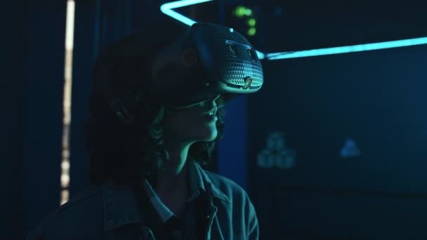 Close-up shot of young woman with curly hair wearing AR headset, standing in dark empty arena with neon lighting, looking around and experiencing augmented reality - Footage, Video