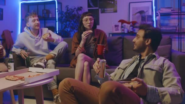 Середній повний знімок молодого арабського чоловіка і кавказької дівчини і хлопця, що сидить разом за столом з пивом і піцою на студентській домашній вечірці, їсть, посміхається і обговорює щось смішне - Кадри, відео