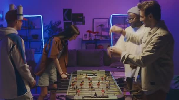 Середній знімок багатоетнічної молоді в повсякденному вбранні, граючи в настільний футбол у двох командах на домашній вечірці в блакитному неоновому вогні, афроамериканський і арабський хлопець виграє і робить п'ять - Кадри, відео