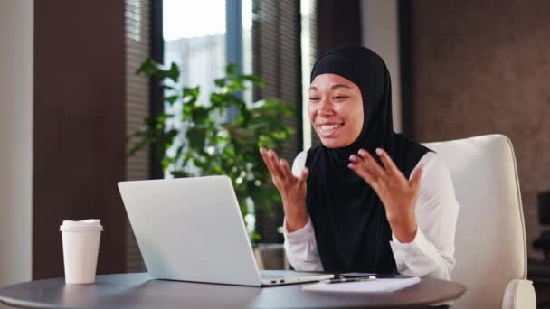 Vrolijke multiraciale kantoormedewerker in zwarte hoofddoek die vreugde uitdrukt op de werkplek. Verrast lachende vrouwelijke gebaren en gebalde vuist in opwinding tijdens het lezen van geweldig nieuws op draadloze laptop. - Video