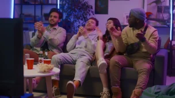 Pełne ujęcie czterech wielonarodowych zoomerów siedzących na kanapie w salonie z fioletowym neonowym światłem, przed telewizorem, z piwem i chipsami, wiwatujących, klaskających i świętujących mistrzostwa swojej drużyny - Materiał filmowy, wideo