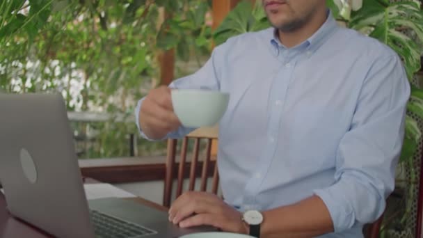 Młody biznesmen w bezprzewodowych słuchawkach, okularach i formalnej koszuli pijący kawę i pracujący na laptopie w kawiarni na świeżym powietrzu w letni dzień - Materiał filmowy, wideo