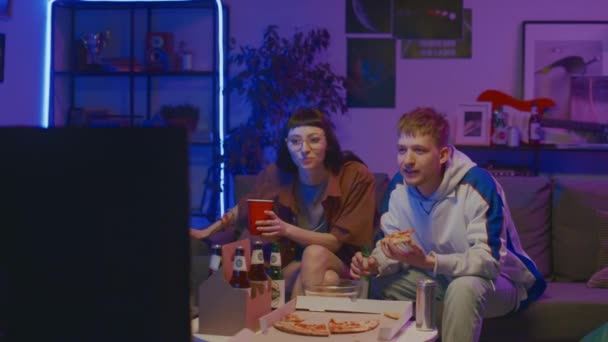 Full shot of young Caucasian couple sitting at home in front of TV on Friday night, eating pizza, drinking beer, watching sport championship, cheering and discussion game - Imágenes, Vídeo