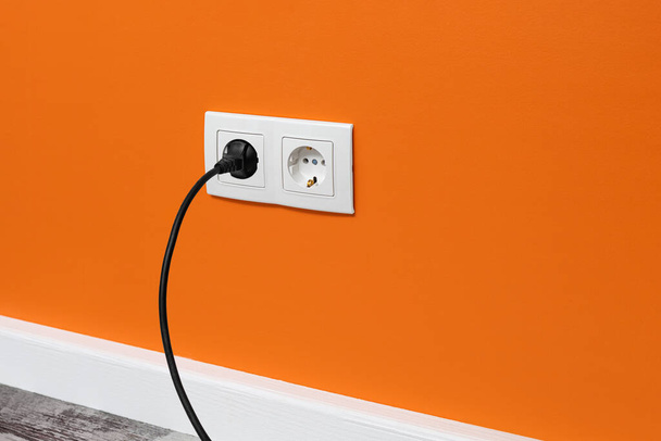 White double outlet installed on orange wall with inserted black electrical plug, side view. - Photo, Image