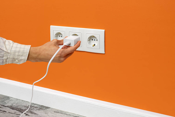 Рука людини підключає телефонний адаптер в потрійній білій електричній розетці, розташованій на помаранчевій стіні, вид збоку. - Фото, зображення