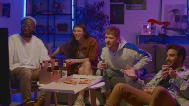 Full shot of young Caucasian girl, African American and Arab guys sitting together on couch at home, eating pizza, drinking beer, watching TV show or sport match, then cheering with joy - Imágenes, Vídeo