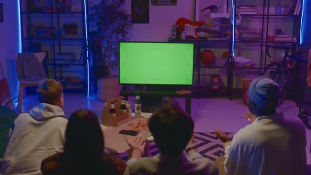 Medium rear shot of four multiracial young people sitting together on couch at home with pizza and beer, watching sport game on TV with empty green screen, cheering and clapping. Template, copy space - Footage, Video
