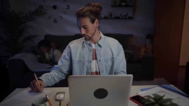 Молодой кавказский мужчина работает сконцентрированным из дома с помощью ноутбука и мобильного телефона. Серьезные люди изучают проект для онлайн бизнеса. Университет серьезный мужчина ищет информацию на компьютере ночью.  - Кадры, видео