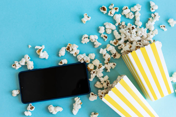 Sárga és fehér csíkos dobozok popcornnal és telefonnal, kék háttérrel. Szórakozás. Filmest popcornnal. Mozi téma. - Fotó, kép