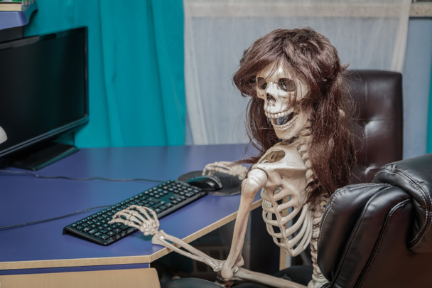 Radosny uśmiechający się szkielet w peruce, siedząc w fotelu za komputer stacjonarny - Zdjęcie, obraz