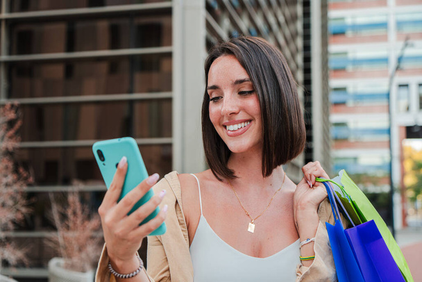 Молодая взрослая радостная женщина, использующая мобильный телефон для покупки он-лайн на рынке приложений. Улыбающаяся женщина с сумками для покупок и весело смотрящая на распродажу по мобильному телефону. Счастливая белая леди. Высокий - Фото, изображение