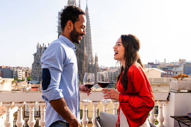 Мультирасовая красивая счастливая пара влюбленных, встречающаяся на балконе на крыше в Саграда Фаза, Барселона - Мультиэтнические люди, имеющие романтический аперитив на террасе с видом на город, понятия о туризме и образе жизни людей - Фото, изображение