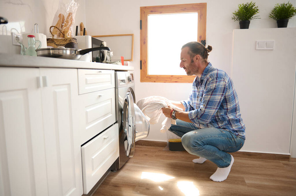 Autentický postranní portrét mladého dospělého vousatého bělocha, který se krčí a nakládá pračku. Je den vyprání. Úklid, domácnost a domácnost. Domácí život. Kopírovat reklamní prostor - Fotografie, Obrázek