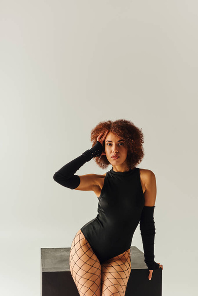 atractiva mujer afroamericana en medias de rejilla y traje de cuerpo sexy posando cerca de cubo negro, moda - Foto, Imagen