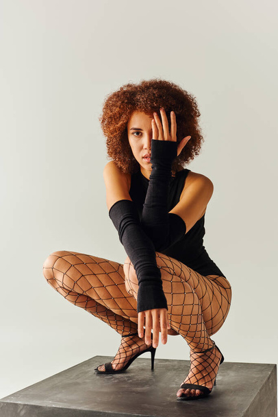 appealing young african american woman in sexy black attire squatting on black cube, fashion concept - Photo, Image