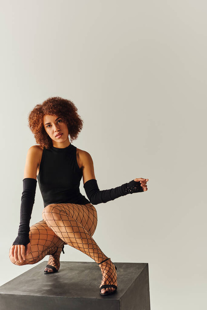 attraente giovane donna afroamericana in sexy abito nero accovacciato sul cubo nero, concetto di moda - Foto, immagini