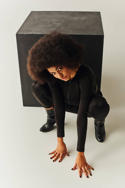 όμορφο αφροαμερικάνικο γυναικείο μοντέλο σε μαύρο bodysuit που ποζάρει δίπλα σε μαύρο κύβο, fashion concept - Φωτογραφία, εικόνα