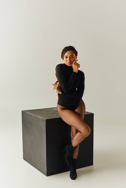 заманчивая африканская американка в сексуальных колготках и боди-костюме позирует рядом с черным кубом, мода - Фото, изображение