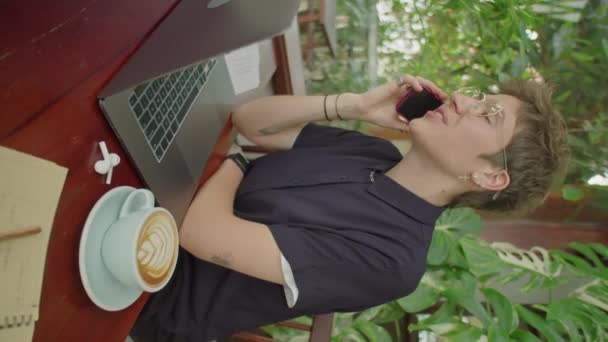 Tiro vertical de jovem empresária de cabelos curtos sentada à mesa com cappuccino e laptop no café ao ar livre e falando no telefone celular - Filmagem, Vídeo