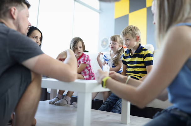 Aikuiset ja lapset istuvat pöydän ympärillä, jossa pelikortit sijaitsevat poika katsoo harkitusti aikuisia. Lautapelejä koko perheen käsite - Valokuva, kuva