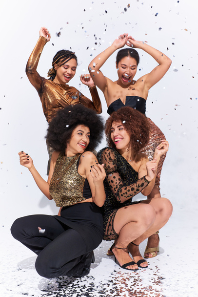 stylische attraktive afrikanisch-amerikanische Frauen feiern unter Konfettiregen auf dem Boden sitzend, Mode - Foto, Bild