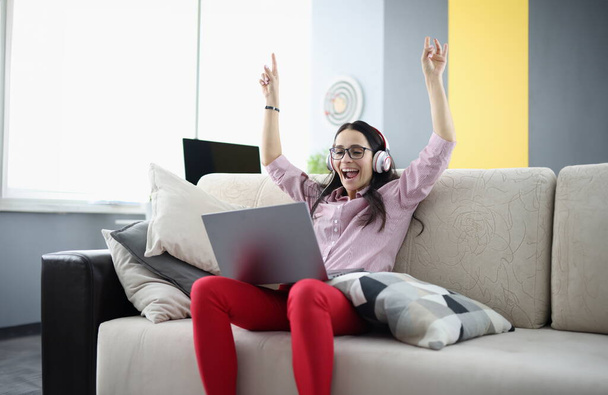 Rejoicing γυναίκα κάθεται στον καναπέ με ακουστικά με φορητό υπολογιστή. Θετική και χαρούμενη στιγμή στο σπίτι έννοια - Φωτογραφία, εικόνα