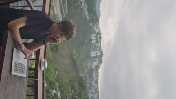 Verticaal schot van jonge zakenvrouw bellen op mobiele telefoon tijdens het werken op afstand op laptop op cafe terras met schilderachtig uitzicht op de bergen - Video