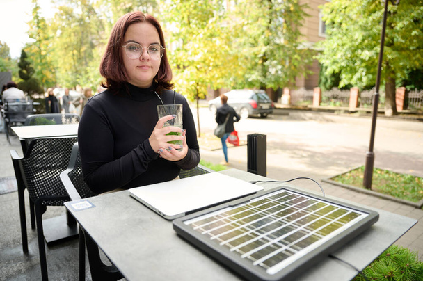 Female using portable solar panel to charge laptop and smartphone. Portrait of a young student girl working remotely from laptop while sitting at a table in a cafe. Drinking mojito. Renewable energy. - Photo, Image
