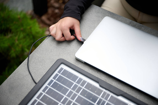 Nahaufnahme eines Mädchens, das einen USB-Stecker in ihren Laptop steckt, um ihn von einem tragbaren Solarpanel aufzuladen. Frauen nutzen tragbare Solarzellen, um Laptop und Smartphone aufzuladen. Erneuerbare Energien. Ausgeschnittenes Bild - Foto, Bild