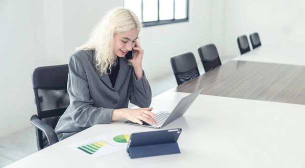 Щаслива молода бізнес-леді використовує ноутбук у конференц-залі всередині офісу Молода жінка-менеджер використовує телефон для розмови з фінансовими та маркетинговими клієнтами. - Фото, зображення