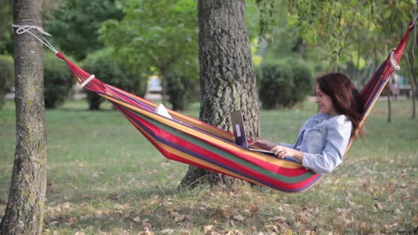Όμορφη νεαρή φοιτήτρια κάθεται σε μια αιώρα και σπουδάζει online με ένα φορητό υπολογιστή στο πάρκο σε μια μέρα του φθινοπώρου. - Πλάνα, βίντεο