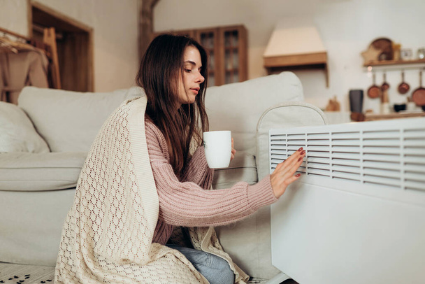 Koud thuis, ijskoud. Het gebruik van verwarming thuis in de winter. Vrouw verwarmt haar handen zittend bij apparaat en draagt warme kleren en houdt warme drank vast. Verwarmingsseizoen. . Hoge kwaliteit foto - Foto, afbeelding