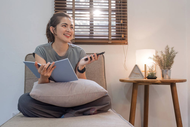 Boldog fiatal nő, aki könyvet olvas otthon a kanapén. Életmód szabadúszó pihenni, és használja okostelefon a nappaliban. Életmód-koncepció. - Fotó, kép