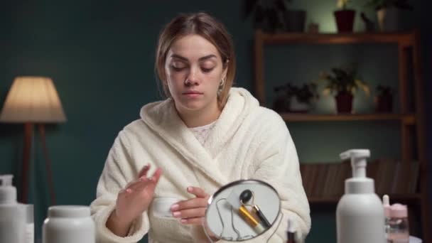 Concepto de cuidado facial. Mujer aplicando crema facial mirando el espejo en casa. Cosméticos femeninos. Copiar espacio - Imágenes, Vídeo