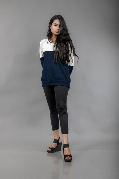 Indiaas jong vrouwelijk model in casual winterkleding tegen grijze achtergrond. Lang zwart harig model met zwarte legging, blauw en wit sweatshirt. - Foto, afbeelding