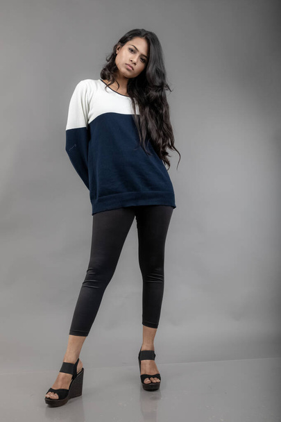 Индийская молодая модель в повседневной зимней одежде на сером фоне. Длинные черные волосы модели в черных леггинсах, синей и белой толстовке. - Фото, изображение