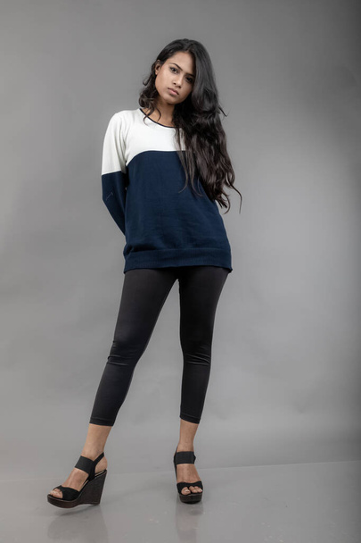 Jeune modèle indien féminin en tenue d'hiver décontractée sur fond gris. Long modèle aux cheveux noirs portant des leggings noirs, sweat-shirt bleu et blanc. - Photo, image