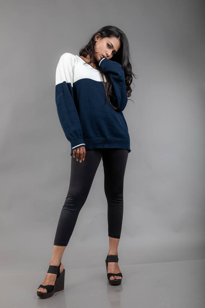Modelo femenino joven indio en ropa de invierno casual sobre fondo gris. Modelo de pelo negro largo con leggins negros, sudadera azul y blanca. - Foto, Imagen