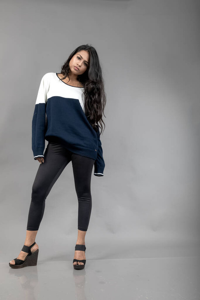 Modelo femenino joven indio en ropa de invierno casual sobre fondo gris. Modelo de pelo negro largo con leggins negros, sudadera azul y blanca. - Foto, Imagen