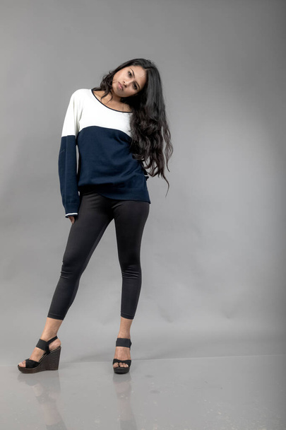 Indian young female model in casual winterwear against grey background. Long black haired model wearing black leggings, blue and white sweatshirt. - Photo, Image