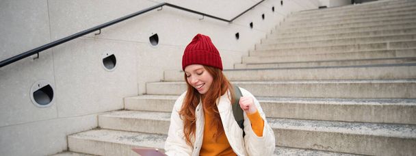 Young beautiful girl with long red hair, smiles, uses internet application, connect to wifi near campus, sits on stairs with digital tablet. - Foto, Imagem