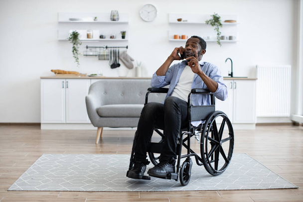 Full length view of adult person in wheelchair talking on cell phone while using comfort of modern studio flat. Mature african man organising birthday party while getting recovery from illness. - Foto, immagini