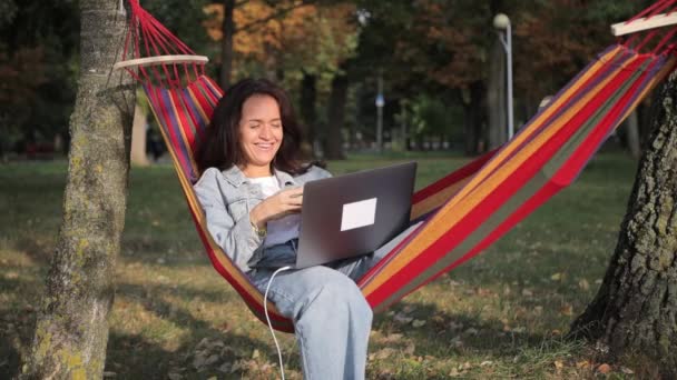 Žena na volné noze ležící v houpací síti v parku pomocí notebooku. Práce na čerstvém vzduchu. - Záběry, video