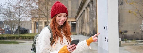 Smiling redhead girl tourist, walks around city and explores popular landmarks, sightseeing, holding smartphone, looking at her mobile app and checks with city map. - Valokuva, kuva